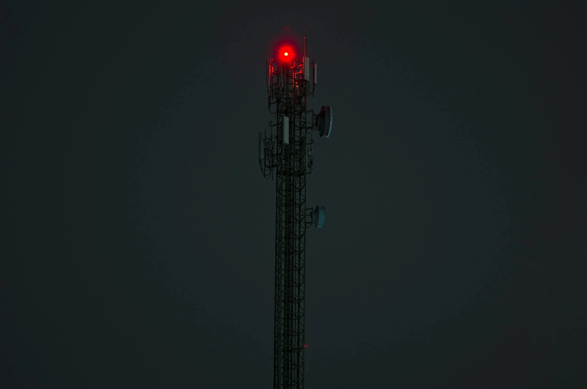 A telecommunications mast at dusk, its warning light lit against a dark sky.